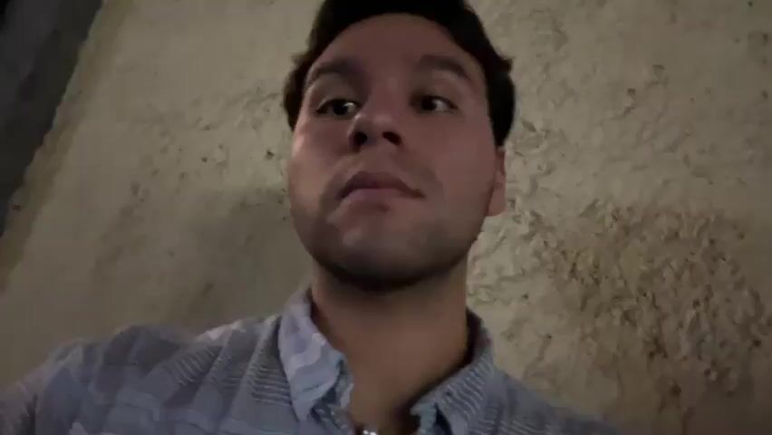 A man wearing a button down shirt is looking at the camera. He looks surprised. In the background are two other men standing outside on a wet street. One is looking at the camera and the other is looking at his phone.