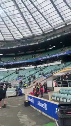 The video shows the interior of Allianz Stadium, a large sports stadium with a green seating arrangement. The camera angle pans across the stadium, capturing the field, the stands with scattered spectators, and a large overhead scoreboard displaying the word "England." There are several people on the field, including groups of individuals who appear to be staff members in black clothing and uniformed personnel in orange vests. A man in a black jacket walks into the frame. The scene is set during a cloudy day, with artificial lighting illuminating the stadium. Allianz signage is visible on the barrier near the camera.