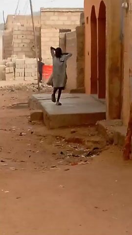 A young person in a gray garment walks away from the camera. The individual has their hands behind their head, and they are standing on a concrete platform. The platform is in a sandy, dusty area. A building with orange or brown columns is to the right of the platform. In the background, there are piles of concrete blocks, and another individual is standing in the distance. There is debris and trash scattered around the ground. A skinny tree trunk stands to the right of the frame.