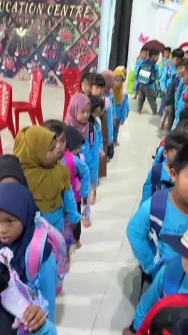 A group of young children, appearing to be students, are lined up indoors. They are all wearing similar light blue long-sleeved shirts and many have blue backpacks. Some of the girls are wearing headscarves in pink and yellow. In the foreground, a young boy with a light-colored baseball cap is looking towards the camera, and another boy with a red backpack is partially visible below him. A red plastic chair is visible to the right of the line of children. The background features a colorful wall decoration with cartoon characters and a rainbow. The overall environment appears to be a school or a similar educational facility. The children seem to be waiting or participating in an activity.