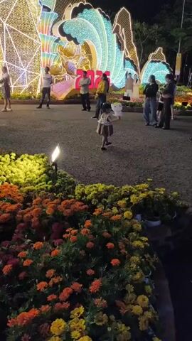 At night, a small child with a dress and pigtails walks in the middle of a paved park area. In front of her is a bright, illuminated art installation with the number 36 on it. Several people are milling around the installation. Some people are taking photos. There are flower beds to the left of the path. The child is carrying a small, white balloon. A woman in a black shirt and white flower patterned pants walks toward the right of the frame.