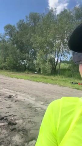 A woman wearing a black baseball cap, black sunglasses, and a neon yellow long-sleeved sheer top over a yellow bikini walks along a sandy and muddy shore. She smiles at the camera and adjusts her sunglasses. In the background, there are trees, a body of water, and several cars. A red and black bag is slung over her shoulder.