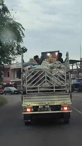 A white Isuzu truck with a metal cargo bed is driving on a paved road under a cloudy sky. The truck is filled with various items, including what appear to be bundles wrapped in white plastic or tarpaulin, and possibly some building materials. Two people are seated in the back of the truck amidst the cargo. One person is wearing a yellow shirt and the other is wearing a blue shirt. A black pickup truck with a ladder is visible in front of the Isuzu truck. To the left of the road, there are buildings with a light pink facade and shops. Power lines are visible overhead. The license plate of the Isuzu truck is partially visible and reads "AL 8148 ZM". The camera is positioned behind and slightly to the right of the Isuzu truck, capturing the scene from a moving vehicle.