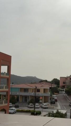 A static shot captures a view of a street lined with buildings and parked cars under a hazy sky. On the left, a partially visible orange building with open balconies dominates the foreground. Across the street, a two-story yellow building with red-tiled roofs features the UniCredit logo and several parked cars in front. To the right of the yellow building, a street curves upwards, with more parked cars visible. Further to the right, a multi-story building with balconies, some enclosed with glass, stands. In the background, a hilly landscape is visible under the overcast sky. The overall scene suggests a quiet residential or commercial area on a cloudy day.