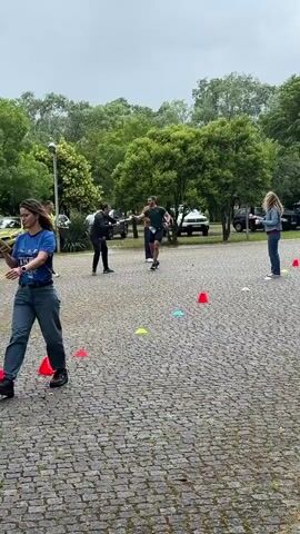 A man is running through a course marked by cones. There is a blue tent nearby. People are standing and watching. The area is paved with cobblestones. Cars are parked in the background. It appears to be a cloudy day. The man drinks from a cup as he runs.