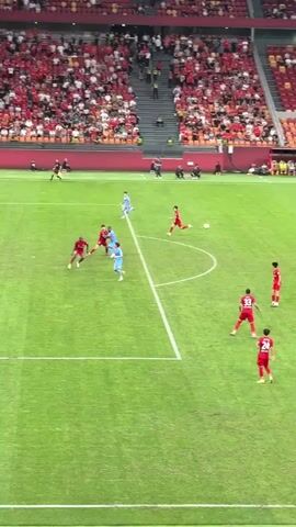 The video shows a soccer game in a stadium with a large crowd. Players in red and blue uniforms are on a green field, and a player in red shoots the ball towards the goal, which is defended by a player in green. The crowd is predominantly dressed in red and waving red flags, and the stands are packed.