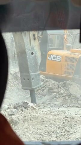 The video captures an excavator with a hydraulic breaker attachment actively breaking apart rocks and debris on a construction site. The excavator is yellow and black with the JCB logo visible. The hydraulic breaker is a large, heavy-duty tool with a chisel-like end. The scene is dusty, suggesting active demolition or excavation. The view appears to be from inside another vehicle, possibly a cabin or truck, looking through a windshield or window. A person's head and shoulder, wearing an orange garment, are visible in the lower left corner, indicating a human presence within the viewing vehicle.