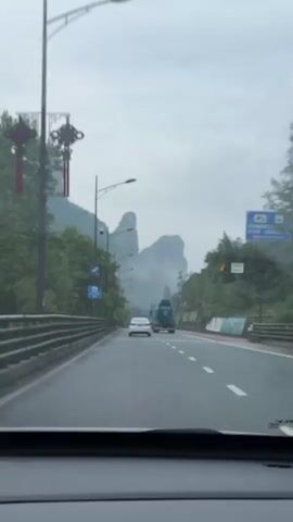 The video shows a car driving down a two-lane road with a concrete barrier to the left and a red-striped shoulder to the right. The road surface is wet. In the distance are tall, rocky mountains obscured by clouds. A white sedan and a blue truck are ahead on the same road. A sign in Chinese is visible on the left, along with street lights and foliage. The camera is positioned inside the car, looking out through the windshield.  The audio includes a warning in Chinese to be cautious of falling rocks.