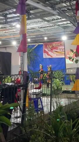 A group of people are gathered under a covered structure, possibly an outdoor venue or event space, with a metal roof and exposed beams. Colorful banners, some triangular and some rectangular, hang from the ceiling in shades of purple, yellow, and red. In the background, a large banner is displayed, featuring the Indonesian flag and text related to the country's anniversary. Two individuals dressed in traditional or ceremonial attire are standing on a raised platform, facing the audience. In the foreground, a metal fence separates the audience from the performance area. Several people are visible behind the fence, some wearing red tops, appearing to be part of the event staff or performers. There are chairs and tables in this area. Lush green plants are growing in front of the fence, adding a natural element to the scene. The lighting suggests it is evening or a dimly lit indoor space, with spotlights illuminating the performance area. The overall atmosphere appears to be festive, possibly a cultural performance or celebration.