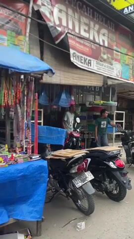 The video shows a street scene in what appears to be a Southeast Asian country. There are several small businesses along the street. One is "ANEKA," which sells household goods, including carpets and gifts. Another is "NABAHOUSE," which sells just-all-brand products. People are walking and driving motorbikes along the street. A person in a green shirt is standing in front of ANEKA. A woman wearing a hijab is riding a motorbike in front of NABAHOUSE. Other motorbikes are parked on the side of the road. The weather looks overcast. The video is shot from inside a moving vehicle.