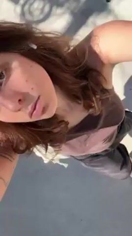 The video shows a person from a high angle looking downwards. The person has shoulder length brown hair and is wearing a brown top, and black shorts, and black sandals. There is a white cloth bag in their hand. The person is walking on a grey concrete surface with various elements, like a drain and a metal fence, appearing in the background.  The camera viewpoint moves along with the person as they walk. There is a shadow of the person visible on the concrete surface.