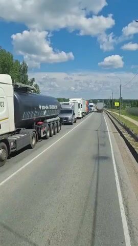 The video shows a traffic jam on a highway under a cloudy blue sky. Several large trucks and some cars are lined up on the asphalt road, indicating a standstill or very slow movement. The trucks appear to be commercial vehicles, with visible company names and logos on some of them. One truck in the foreground is a tanker with Cyrillic text. Another truck has a white trailer with blue lettering that appears to be "Turbo Trans". A bright orange truck with a blue container is also visible further down the line. The surrounding environment includes green trees and grass on the side of the road, with a fence and a yellow warning sign. The perspective is from a vehicle driving behind the traffic jam, suggesting the video was recorded by a passenger or driver.