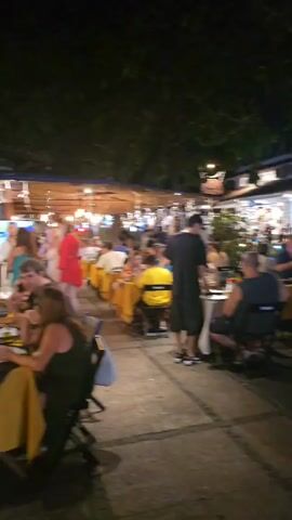 The video shows a lively outdoor dining scene at night. Numerous people are seated at tables with yellow tablecloths, while others are walking around. A waiter is attending to a table. A food cart with bright lights attracts customers. The scene is lit by artificial lights and street lights. Trees surround the area, creating a pleasant ambiance.