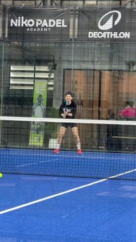 A woman in a black t-shirt and black shorts, with pink socks and red shoes, plays padel. She is on a blue court with a net. She is holding a padel racket and hitting a yellow ball. The background shows a glass wall with logos for "NIKO PADEL ACADEMY" and "DECATHLON". There are other people in the background, some watching and some playing. The lighting is artificial, suggesting an indoor facility.