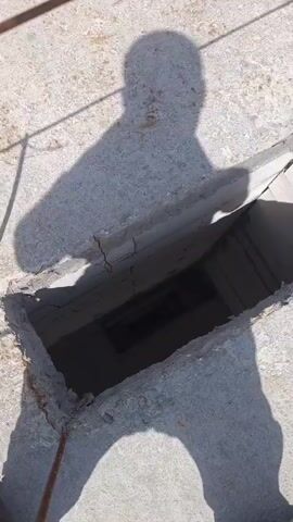 The video pans down into a rectangular opening in a concrete surface. The opening appears to be a shaft or well. The walls of the shaft are made of concrete blocks. At the bottom of the shaft, there is a dark rectangular object, possibly a door or grate. The lighting in the shaft is dim. The overall impression is of an abandoned or disused space.