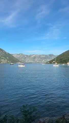 A vertical, medium-shot video shows a wide expanse of blue water with small waves, reflecting a clear blue sky with scattered white clouds. In the distance, a range of majestic, rocky mountains forms a dramatic backdrop, their slopes partially covered in green vegetation. Several white sailboats are anchored or drifting on the water, their masts standing tall against the sky. One sailboat in the foreground is particularly prominent, with people visible on its deck. Other boats, including a larger white vessel, are further out on the water. The overall scene conveys a sense of tranquility and natural beauty on a sunny day.