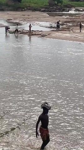 The video captures a wide shot of a muddy river and its sandy banks. Several people are visible on the bank, some appearing to be working or interacting in small groups. In the foreground, a young person walks through the shallow water, carrying an object on their head. The water appears somewhat murky, with ripples indicating a gentle flow. The overall scene suggests an outdoor environment, possibly a rural or developing area, where people are engaged in activities by the river.