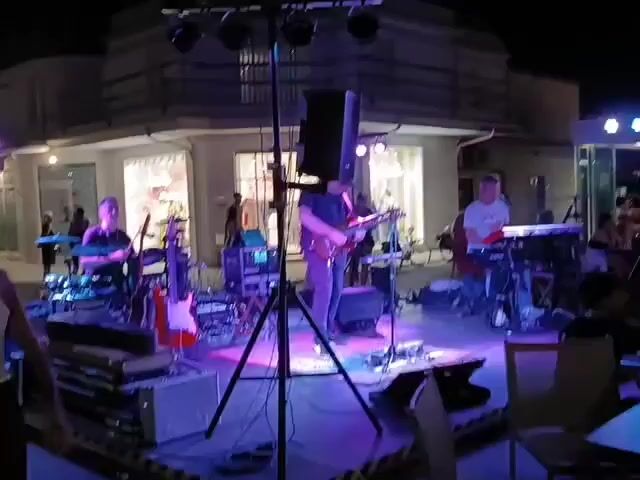 A band is playing on an outdoor stage at night. The stage is lit with blue and purple lights. A drummer is on the left, playing drums. A guitarist is in the center, playing a guitar. Another musician is on the right, playing a keyboard. There are several speakers and other musical equipment on the stage. In the background, there is a building with windows. There are a few people standing in front of the building. The overall scene suggests a live musical performance.