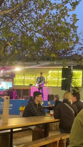 The video shows a street concert with a guitarist playing on a stage. The guitarist, dressed in a denim vest and jeans, plays with a focused expression. The stage is decorated with yellow lights and a white wooden facade. In the foreground, people sit at picnic tables, enjoying drinks and watching the performance. The scene takes place outdoors, under a tree with sparse foliage, at dusk.