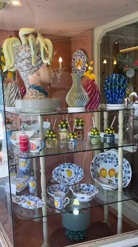 The video shows a display case filled with decorative items, primarily ceramics, in a store. The display case is made of glass and has multiple shelves. The items on display are colorful and feature various designs, including what appears to be a bust with octopus tentacles for hair, vases in different shapes and colors, plates, bowls, mugs, and diffusers with lemon motifs. The lighting in the store is visible, with a spotlight illuminating some of the items on the bottom shelf. The background is slightly out of focus but appears to be a retail environment. No people are visible in the video, and no actions or interactions are occurring. The overall scene is static and focused on the merchandise.