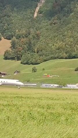 A white jet airplane is moving across a grassy field towards a runway. The airplane is moving from left to right in the frame. In the background, there are buildings and a forest-covered hill. There are also two small red helicopters visible on the grass field in the middle distance. The video is a close-up of the airplane.