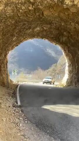 The video shows a grey SUV driving through a short tunnel. The tunnel is made of rough, light brown rock. The road is asphalt and has a white stripe on the left. Outside the tunnel, there are trees and mountains in the distance. There are also road signs on the left. The car is moving towards the camera. The sun is shining.