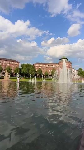 A wide shot shows a large fountain with water spraying upwards in a body of water in the foreground. In the background, there are buildings and trees. A person wearing a white shirt and dark pants is visible near the fountain in the background. There are also several statues or sculptures visible in the background. The sky is blue with white clouds.