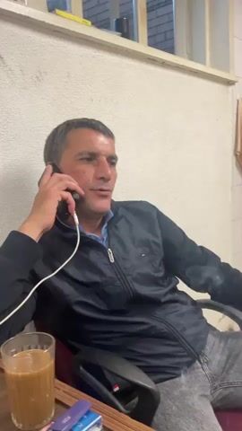 A man with short, graying hair is sitting in a black office chair and talking on a black smartphone held to his left ear. He is wearing a dark blue zip-up jacket over a blue collared shirt and gray jeans. He has a cigarette in his right hand, which is brought up to his mouth. In the foreground, on a wooden table, is a glass of brown liquid, possibly coffee, and a few vape pens. The background shows a white wall and a window with blinds.