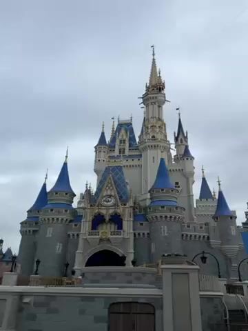 The video shows a view of Cinderella's Castle in Disney World on a cloudy day. The camera pans from the front of the castle to a side turret, showcasing the castle's architecture and the surrounding decorative elements, like ornate street lamps and banners.  The weather appears overcast.