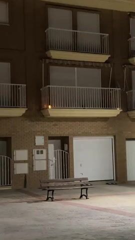 The video shows a three-story building at night. The building is brown with white balconies and closed blinds on the windows. There are garage doors on the ground level. A bench sits in the paved area in front of the building, and a street light illuminates the scene. There is no one visible in the video, and no actions are observed.