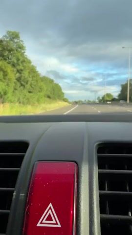 The video shows a close-up view of a car's dashboard, specifically the hazard light button, with the road and landscape visible through the windshield. The hazard light button is red with a white triangle symbol, indicating an emergency or warning. The scene outside the car depicts a highway lined with lush green trees and grass on the left, with a clear sky overhead, though some clouds are present. Streetlights and other vehicles can be seen in the distance on the highway, suggesting a relatively clear and open road. The time of day appears to be late afternoon or early evening, given the lighting and the sky conditions. The location context provided, Malvern Hills, United Kingdom, suggests this is likely a scenic drive in the British countryside. There are no people visible in the frame. The activity is the car driving on a highway.
