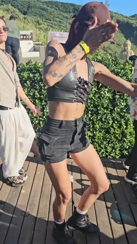 A woman is dancing outdoors on a wooden deck, on a sunny day. She is wearing a brown baseball cap, a black leather-look halter top with a laced front, black shorts, white socks, and black boots. The woman has tattoos on her arms and legs. A man wearing a Hawaiian shirt and black jeans stands to her right, while another man in a patterned shirt and light-colored pants is to her left. A lush green hedge is behind the woman. A hill with some people is in the background. White bits of trash are on the deck near the woman's feet. Other people are mingling in the background.