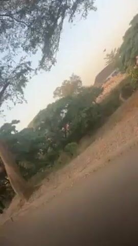 The video shows a street scene during daylight. Two people are walking along the side of the road, near trees and shrubbery. One individual wears a red shirt and the other wears a white shirt. The camera appears to be moving, possibly from a vehicle. The sky is visible through the trees.