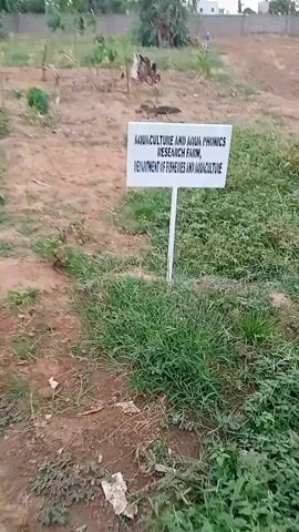 The video shows a sign in front of a field. The sign reads "AQUACULTURE AND AQUA PHONICS RESEARCH FARM, DEPARTMENT OF FISHERIES AND AQUACULTURE".  The field appears to be a mix of bare ground and some grass. In the background, there are trees and what appears to be a building or structure. The video is a little blurry.