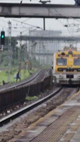 A yellow and white train approaches a station platform on a cloudy day. The train has the number 5053 and "ICF" printed on its front. A person wearing a dark shirt and pants is walking along the train tracks, carrying a blue object. Another person is partially visible near the train's entrance. Overhead, electrical wires crisscross the sky. The platform is paved with yellow and gray tiles.