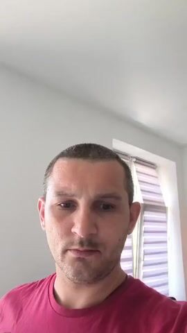 The video shows a close-up, medium shot of a man's face. He has short, dark hair, light skin, and is wearing a red t-shirt. His expression is neutral, with his eyes looking slightly down. The background is blurred, but appears to be an indoor setting with light-colored walls and a window with blinds. The lighting is bright, suggesting daylight. There are no other people or significant objects visible in the frame. The man's head is tilted slightly.