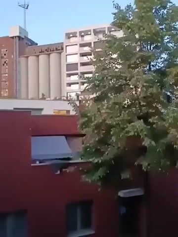 The video shows an exterior view of several buildings, likely in an urban environment. In the foreground, there is a red building with visible windows and balconies. To the left, some green foliage is present. Behind the red building, two taller structures are visible. One is a light-colored, multi-story building with numerous balconies. Adjacent to it is another building with a distinctive industrial design, featuring several large, cylindrical silos and a taller, brick-like section topped with an antenna. The sky appears to be a light blue, suggesting it might be daytime, possibly early morning or late afternoon. The camera pans slightly to the right, capturing different angles of the buildings and the tree.