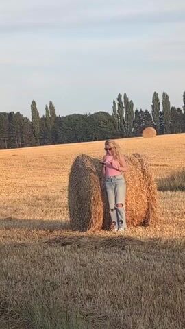 A young woman with blonde, curly hair, wearing a pink tank top and distressed denim jeans, stands in a field of dry hay. She is holding a smartphone and appears to be taking a picture or video of herself. She is positioned in front of two large, round hay bales. The field stretches out behind her, with a line of trees forming the horizon. The sky is a pale blue with a hint of clouds. The overall lighting suggests it is late afternoon or early evening.