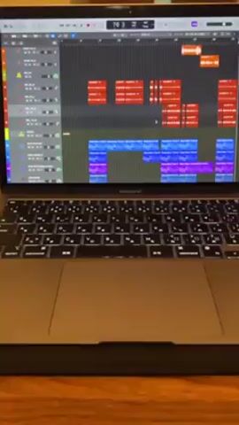 A person is shown pressing the spacebar on a Macbook Air, causing music production software to play a track. The screen of the laptop displays the timeline of the music project, showing different colored blocks representing different tracks. The laptop is on a wooden table.