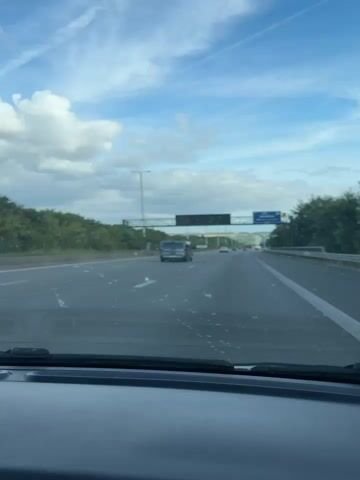 The video shows a first-person view from inside a vehicle traveling on a highway. The road appears to be asphalt with white lane markings. Several cars are visible on the highway, some in front and some in the distance. On the left side of the road, there is a dense green tree line with some power lines and poles visible. On the right side, there is a concrete barrier and more trees.

At approximately 00:01, a large digital sign is visible above the highway. At around 00:02, a road sign with the word "Bouge" is clearly visible on the left side of the road. The sky is blue with scattered white clouds. The overall impression is of a daytime drive on a relatively clear highway.