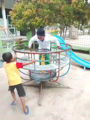 A young boy wearing a yellow shirt, black shorts, and blue sandals is pushing a small merry-go-round at a playground. A slightly older boy dressed in a white shirt, green pants, a black cap, and red shoes stands inside the merry-go-round, holding onto the railing. The merry-go-round is constructed with metal bars and has a tire underneath. The playground setting includes green grass, trees, a blue slide in the background, and parts of other playground equipment visible. The concrete ground shows some wear and tear. The camera angle is static, capturing the scene from a medium distance.