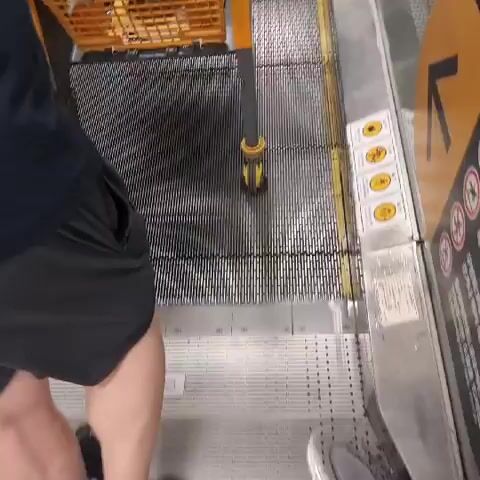 The video shows a person from the waist down, wearing black shorts and a black t-shirt, standing on an escalator and pushing a yellow shopping cart. The escalator steps are gray and ribbed. To the right of the escalator, there is a silver railing with yellow safety markings. Further to the right, there are several circular yellow signs with black icons. The escalator ascends and a few other people are visible further up the escalator, also with shopping carts. The environment appears to be a store with bright lighting.