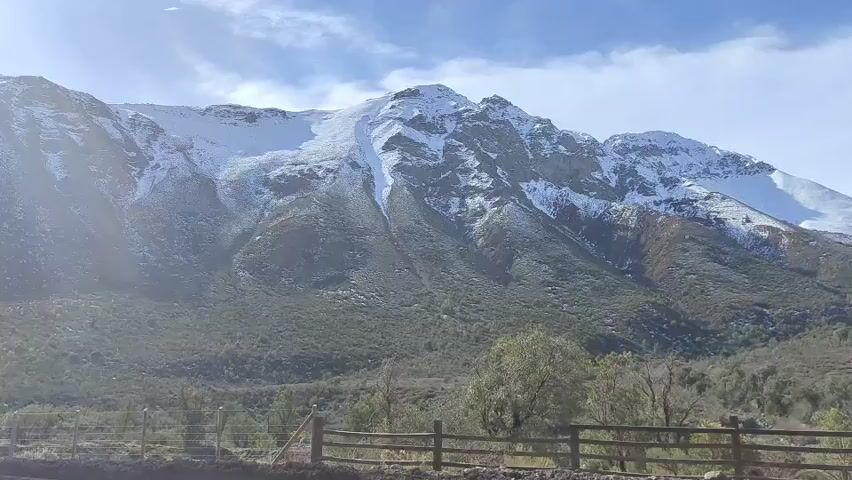 A handheld video shows a mountain range with snow on its peaks. The mountain slopes are covered with trees and shrubs, and there is a wooden fence in the foreground. The sky is blue with some clouds. The video pans slowly from left to right, revealing more of the mountain range.