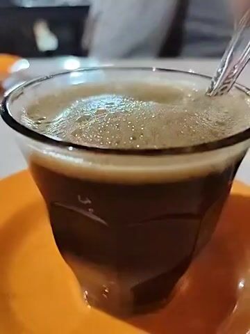 A video shows a hand stirring a cup of dark coffee with a spoon. The coffee is foamy and sits in a faceted glass on an orange plate. The background is blurry, showing a glimpse of what appears to be a table or counter.