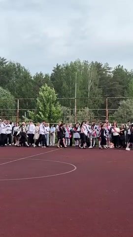 The video shows a group of high school graduates standing on a red outdoor court. They appear to be lined up, likely for a graduation ceremony. Most of the students are dressed formally in white shirts and dark pants or skirts. Some wear sashes with the word "Graduate" in Russian.  They are walking, and some are shaking hands as they pass. A few adults, including what appears to be a teacher, are standing along the line as well. A metal fence runs along the edge of the court, and a forest is visible in the background. The sky is overcast.
