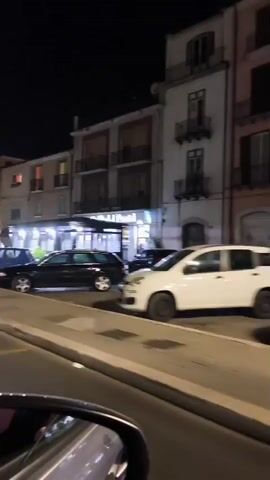 The video shows a nighttime view from a moving vehicle looking out at a street scene. Buildings line the right side of the street, illuminated by interior lights and streetlights. Several cars are parked along the street and some are driving. Yellow lines are painted on the road, including a "BUS" marking. There appear to be a few people standing on the sidewalk. The atmosphere suggests an urban or town setting at night.