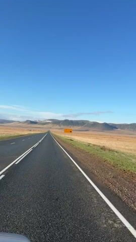 The video presents a view from what appears to be a car moving on a two-lane paved road through a wide, open landscape. The sky is a clear, bright blue. On either side of the road are expanses of fields, transitioning from green to a dry, golden color. In the distance, a range of low mountains is visible, some with snow-capped peaks. A yellow road sign stands along the roadside. The video appears to be taken during the daytime in sunny weather. There is strong wind.