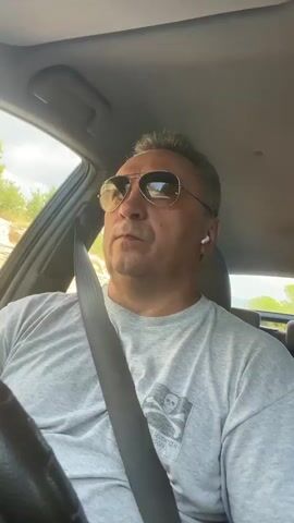 A man in sunglasses and a grey t-shirt with a graphic on it, wearing a seatbelt and earbuds, is driving a car. He looks around outside the car and appears to be speaking. The interior of the car is visible, including the steering wheel and dashboard. The exterior view shows trees and a road. The lighting suggests it is daytime.