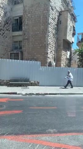 This media file captures a street scene in al-Eizariya, Palestinian Territories. The focus is on a sidewalk and the adjacent buildings, with a man walking along the sidewalk.

The **subjects** include a man dressed in a white shirt and dark pants, wearing a white cap. He appears to be walking and holding something in his hand, possibly a phone or a small bag. The **scene** is dominated by a tall, light-colored stone building on the left and a similar building on the right, with a large tree providing shade in the middle. In the foreground, a street with visible red markings is present. A white metal fence runs along the sidewalk, separating it from the buildings.

The **activity** is simply a man walking along the street. The **setting** appears to be an urban or semi-urban environment, with older stone architecture and modern elements like air conditioning units visible on the buildings. The **time of day** is clearly daytime, indicated by the bright sunlight and strong shadows cast by the tree. The **weather** appears to be sunny and warm, as suggested by the clear blue sky and the light attire of the man.

There is no **visible text** in the media file. The overall **emotion** conveyed is neutral, depicting a commonplace street scene. The location cues like the architecture and the presence of a fence suggest a Middle Eastern context, consistent with the provided location.