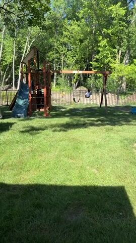A young child is swinging on a swing set in a backyard. The child is wearing a t-shirt with a shark pattern. The swing set is made of wood and has two swings. There is a slide attached to a wooden play structure. The yard is covered in grass and surrounded by trees. A fence separates the yard from a wooded area.